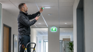 Commercial electrician testing a ceiling-mounted emergency light in a modern office corridor.
